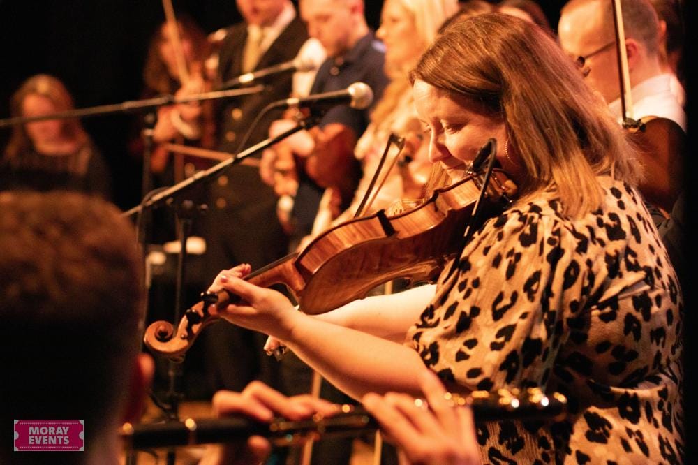 Moray Events, live musical performance with a woman playing the violin at an indoor concert, featuring an orchestra, microphones, and an enthusiastic audience.