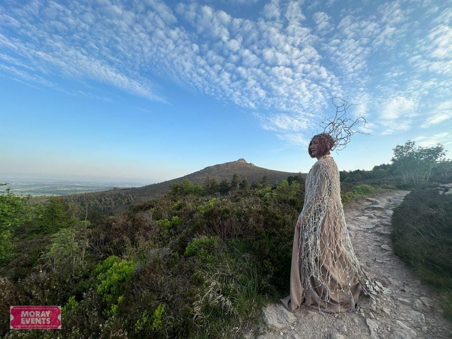 Vibrant outdoor landscape with a woman in a unique, artistic costume on a mountain trail under a blue sky with scattered clouds, showcasing nature-themed costume design for Moray Events.