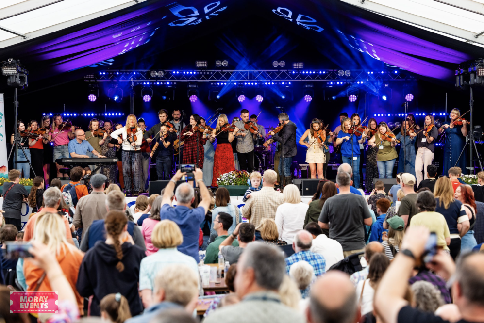 Live outdoor concert with a large audience enjoying a performance by an orchestra under a tent. The event showcases Moray Events' vibrant festivals and community gatherings.