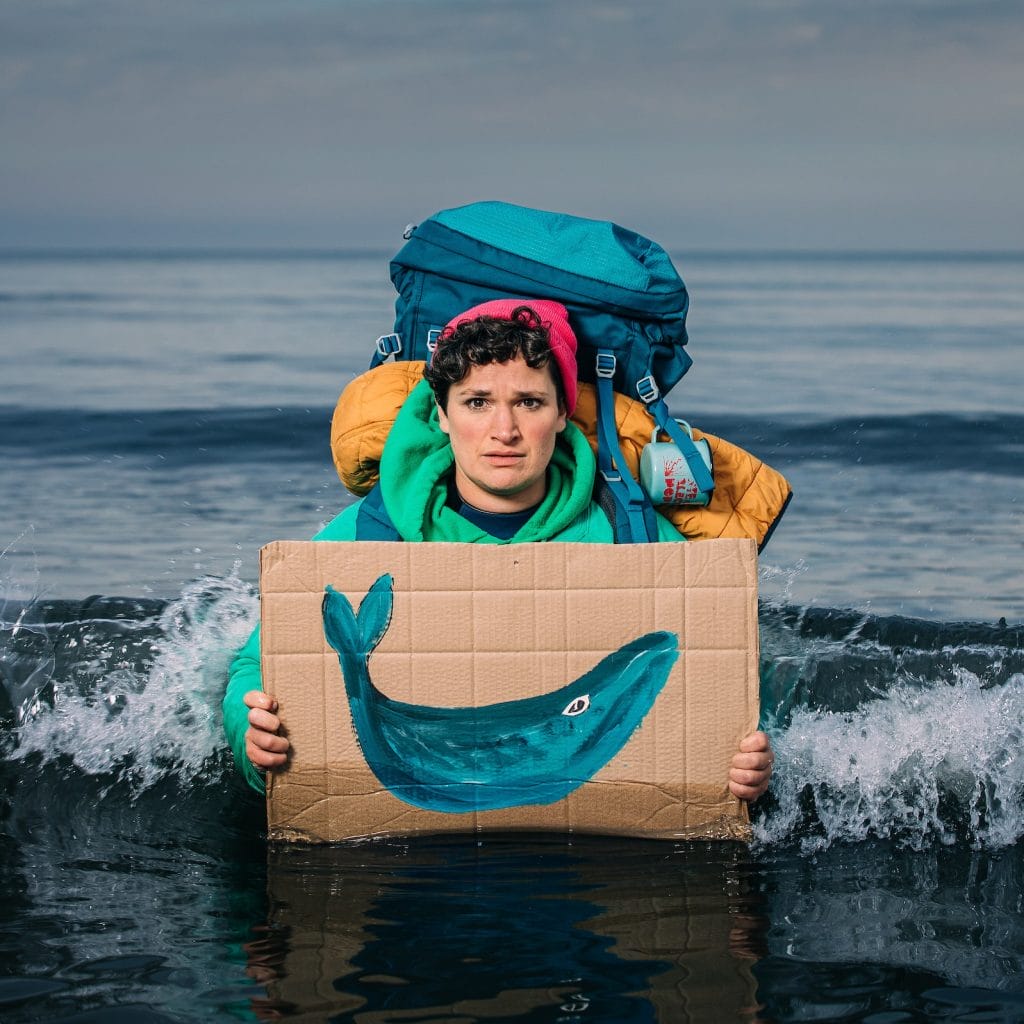 A person with a large backpack holding a cardboard sign with a painted whale in water, symbolising environmental awareness and ocean conservation, on a stormy sea background.