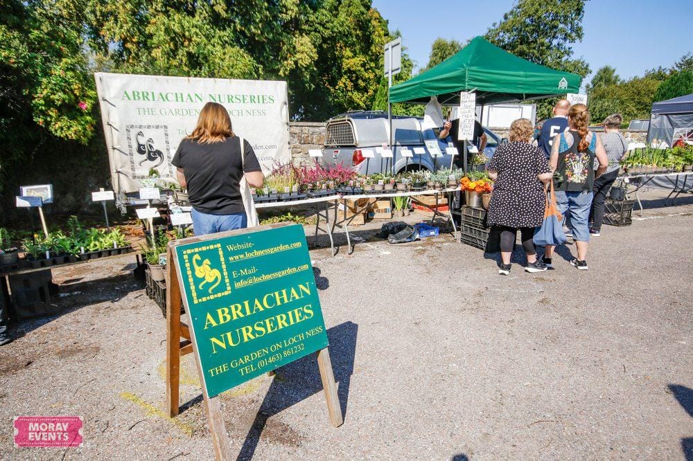 Colourful nursery stall at Moray Events outdoor market featuring a sign for Abriachan Nurseries selling plants and garden accessories. The market has several vendors and visitors enjoying the event.