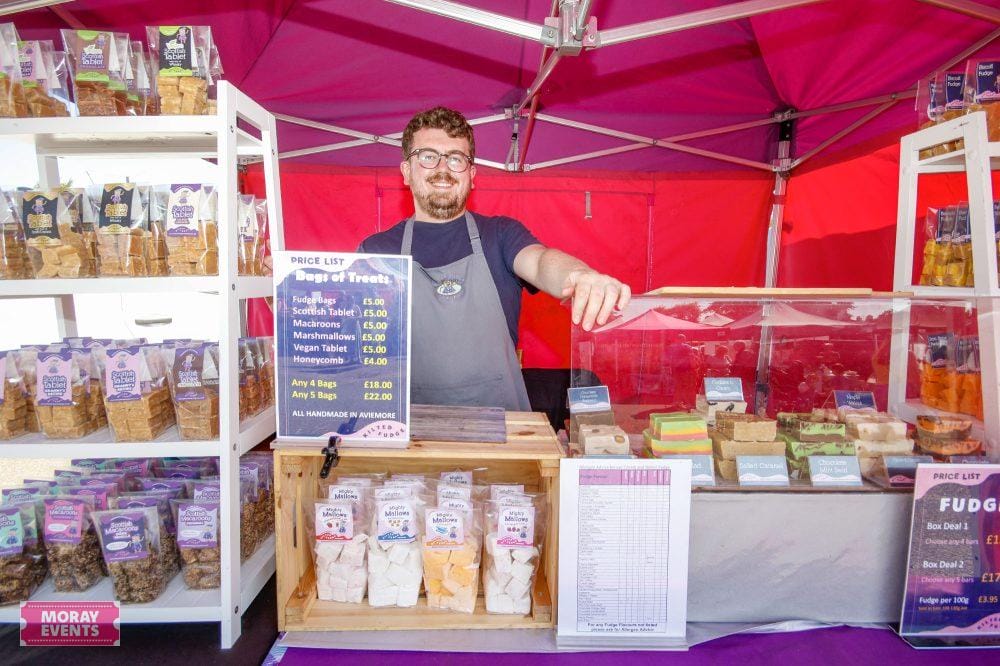 Delicious handmade vegan fudge at Moray Events fair, displayed in colourful blocks behind the stall, with a friendly vendor ready to serve customers under a vibrant pink canopy.