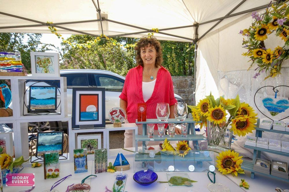 Colourful arts and crafts stall at Moray Events outdoor market, featuring vibrant paintings, handcrafted jewellery, and floral arrangements, under a sunny sky.