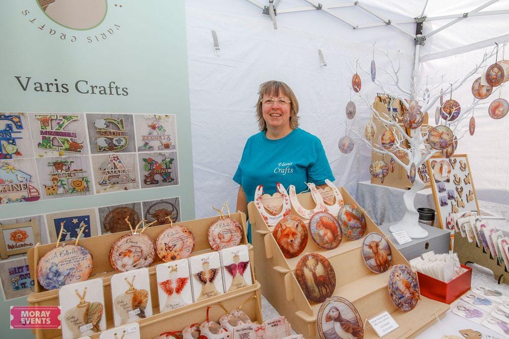 Detailed handmade craft ornaments with animal and nature themes at Moray Events craft fair, featuring a smiling vendor showcasing decorated decorative baubles and ornaments for sale.